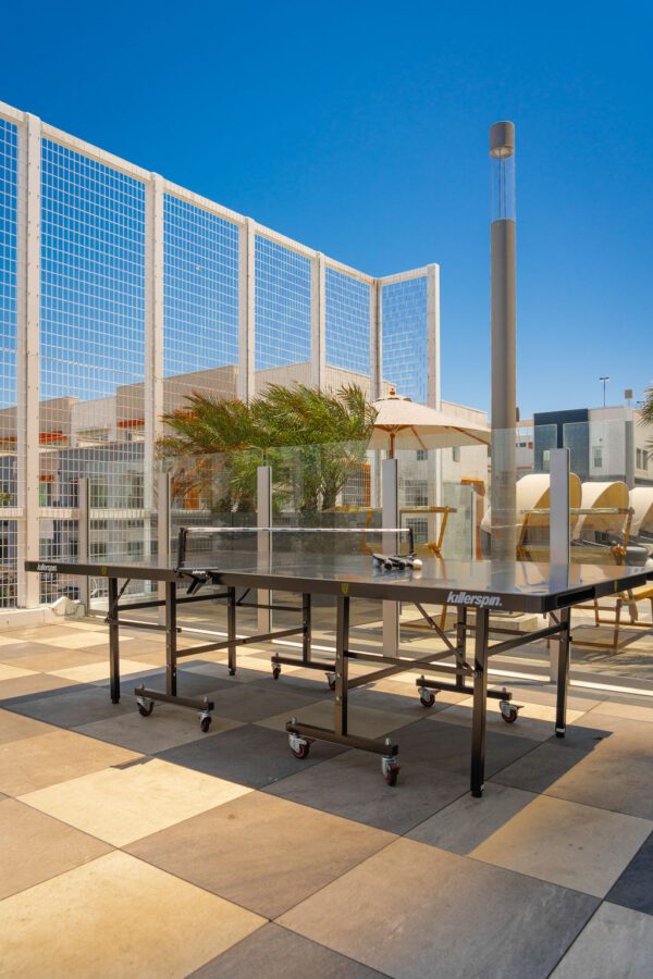 A black ping pong table sits on a tiled rooftop patio with glass railings, lounge chairs, potted plants, and a blue sky overhead.