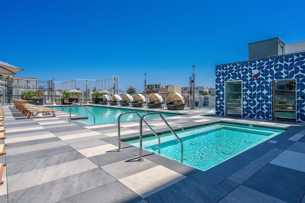 Rooftop pool area with modern lounge chairs, cabanas, and a hot tub under a clear blue sky. The pool deck features geometric tile flooring and a building with a striking blue-and-white patterned wall.