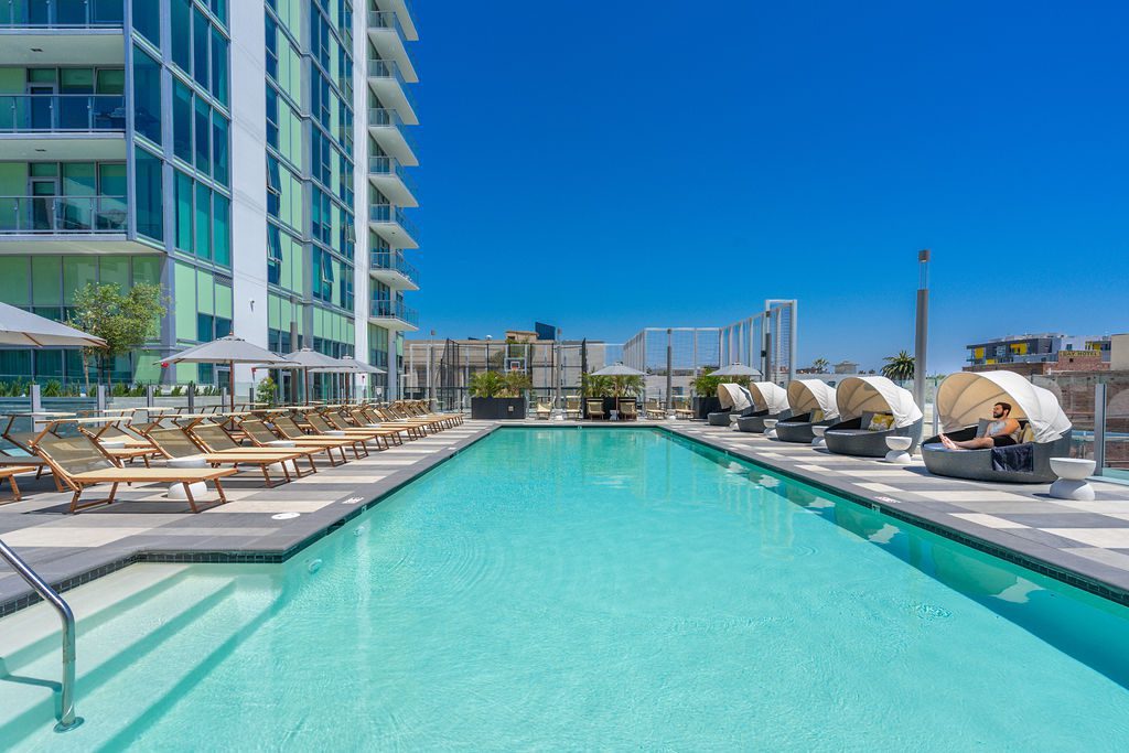 A modern outdoor swimming pool with lounge chairs and shaded cabanas beside a tall building, under a clear blue sky on a sunny day.