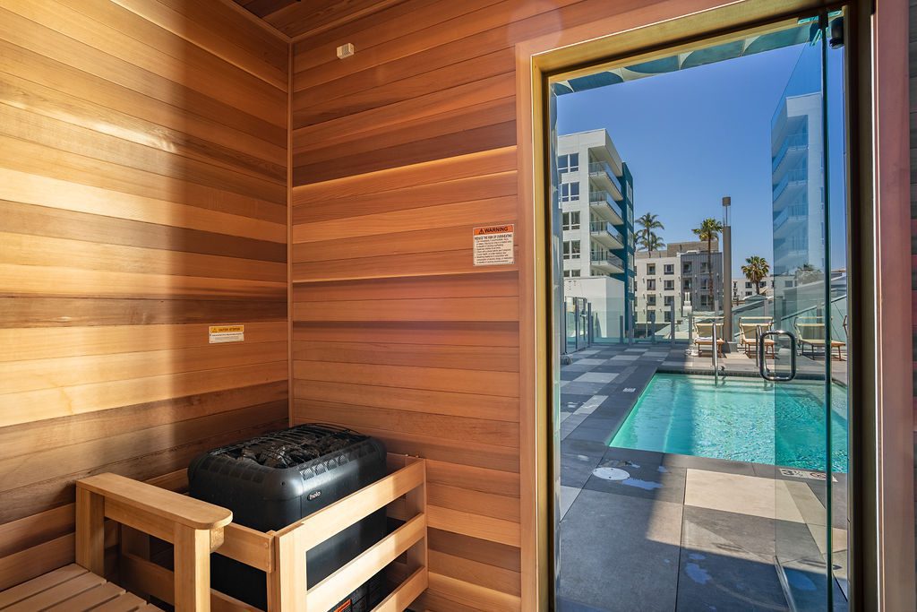 Interior of a wooden sauna with a bench and heater, looking out through a glass door to an outdoor pool area with modern apartments, palm trees, and blue sky in the background.