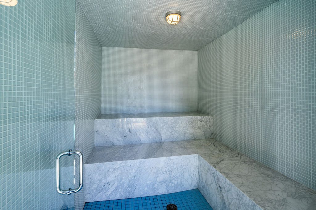 A steam room with light blue mosaic tile walls, marble bench seating, a ceiling light, and a glass door. The floor is tiled in darker blue. The space is empty and clean.