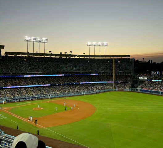 A baseball game at a stadium during sunset, with bright lights illuminating the field, players on the diamond, and the stands filled with spectators. The sky transitions from blue to orange at the horizon.