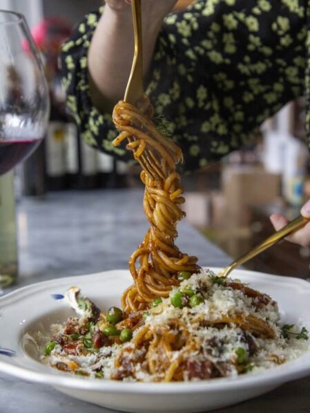 A person twirls spaghetti with tomato sauce, peas, and grated cheese on a fork above a white plate; a glass of red wine and bottles are visible in the background.