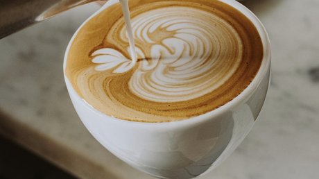 A close-up of a cup of cappuccino as steamed milk is being poured, creating a detailed latte art pattern on the frothy surface.