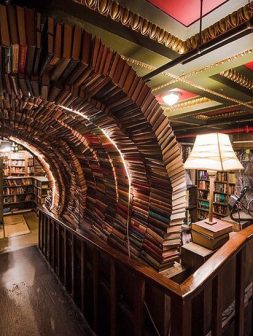 A cozy bookstore features a unique arched tunnel made from stacked books, illuminated by warm lights. Nearby, a vintage lamp sits on a wooden railing, with shelves of books visible in the background.