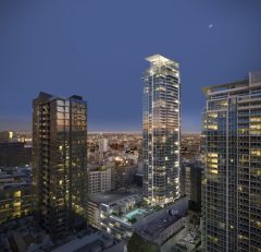 A nighttime cityscape featuring modern high-rise buildings with illuminated windows, a rooftop pool, and a distant view of city lights under a dark blue sky with a crescent moon.