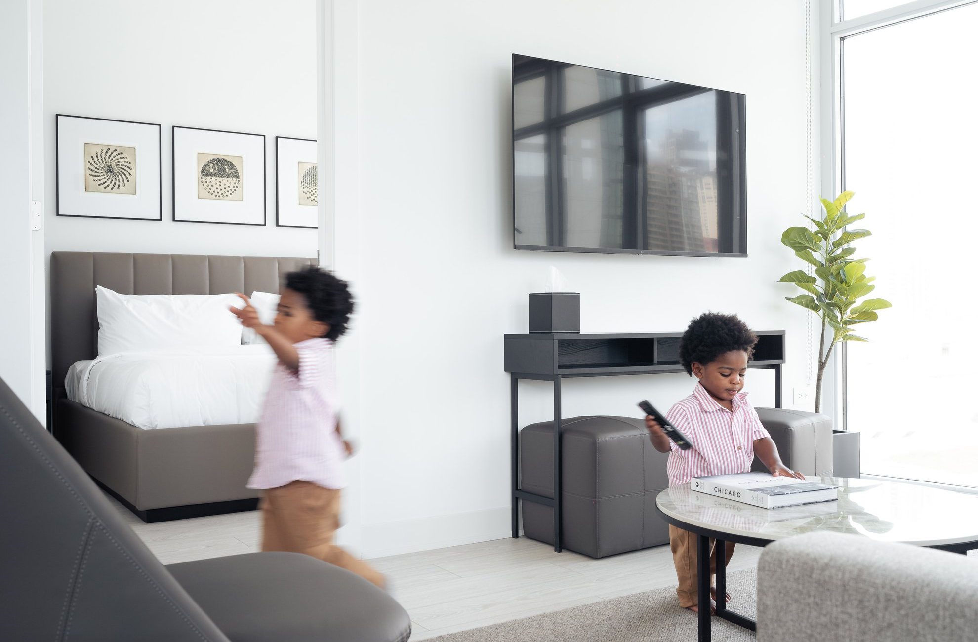 Two young children with curly hair are in a bright, modern living space. One child sits at a round glass table holding a TV remote, while the other walks past a bed in an adjacent room. The area is decorated with art and plants.