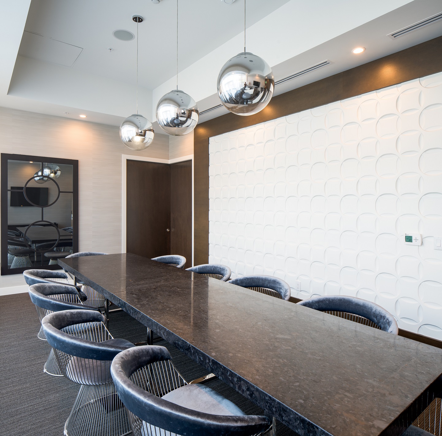 Modern conference room with a long black marble table, eight blue velvet chairs, three metallic pendant lights, a white textured accent wall, and a large mirror reflecting the room.