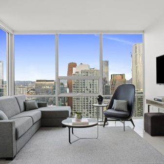 Modern living room with gray sofa, armchair, coffee table, TV, and large windows displaying a cityscape with tall buildings under a clear blue sky.