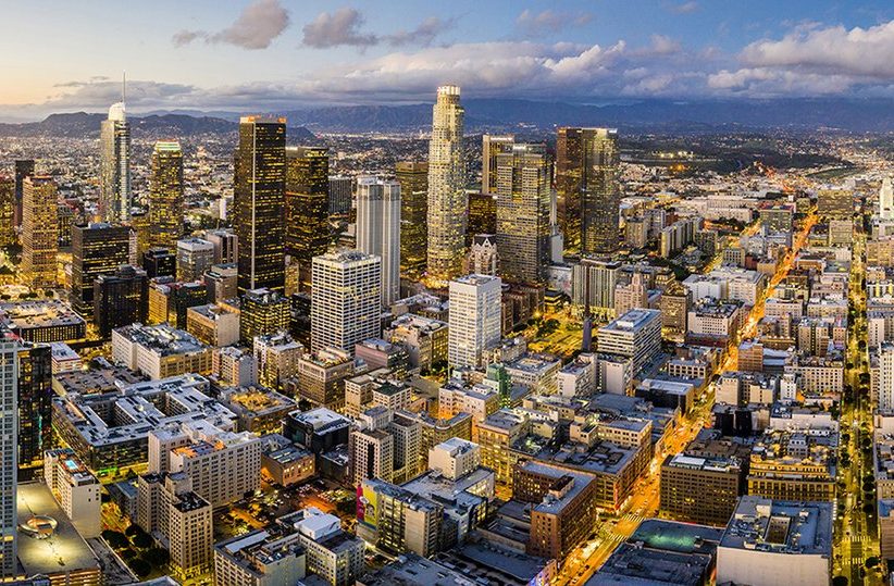 Aerial view of downtown Los Angeles at dusk, showcasing tall skyscrapers, city lights, and a grid of streets with mountains visible in the background under a partly cloudy sky.