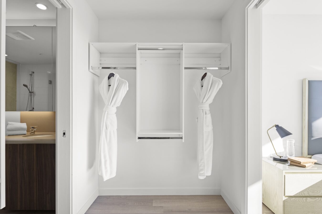 Open closet with two white bathrobes hanging on hooks, shelves above, and no other items. The closet is between a bathroom with towels visible and a bedroom with a nightstand, lamp, and glass of water.