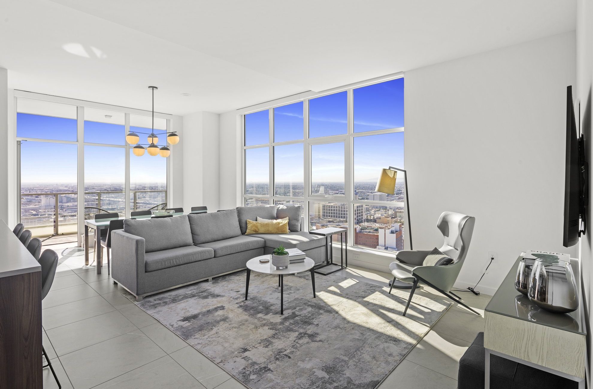 Modern living room with large floor-to-ceiling windows showing a city view, a gray sofa, white armchair, coffee table, dining area, TV on the wall, and plenty of natural light.