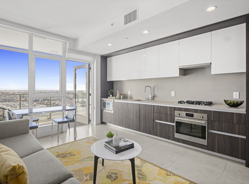 Modern apartment with open kitchen and living area, featuring sleek cabinets, built-in appliances, a gray sofa, round coffee table, and large windows with a view of the city and balcony.