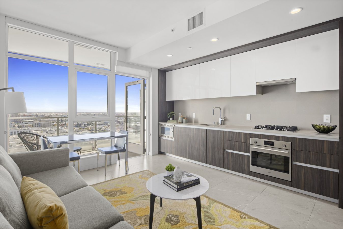 Modern apartment interior with a sleek kitchen, dining table, and sofa. Large windows and a glass door open to a balcony with city views under a clear blue sky. A yellow-patterned rug adds color to the neutral decor.