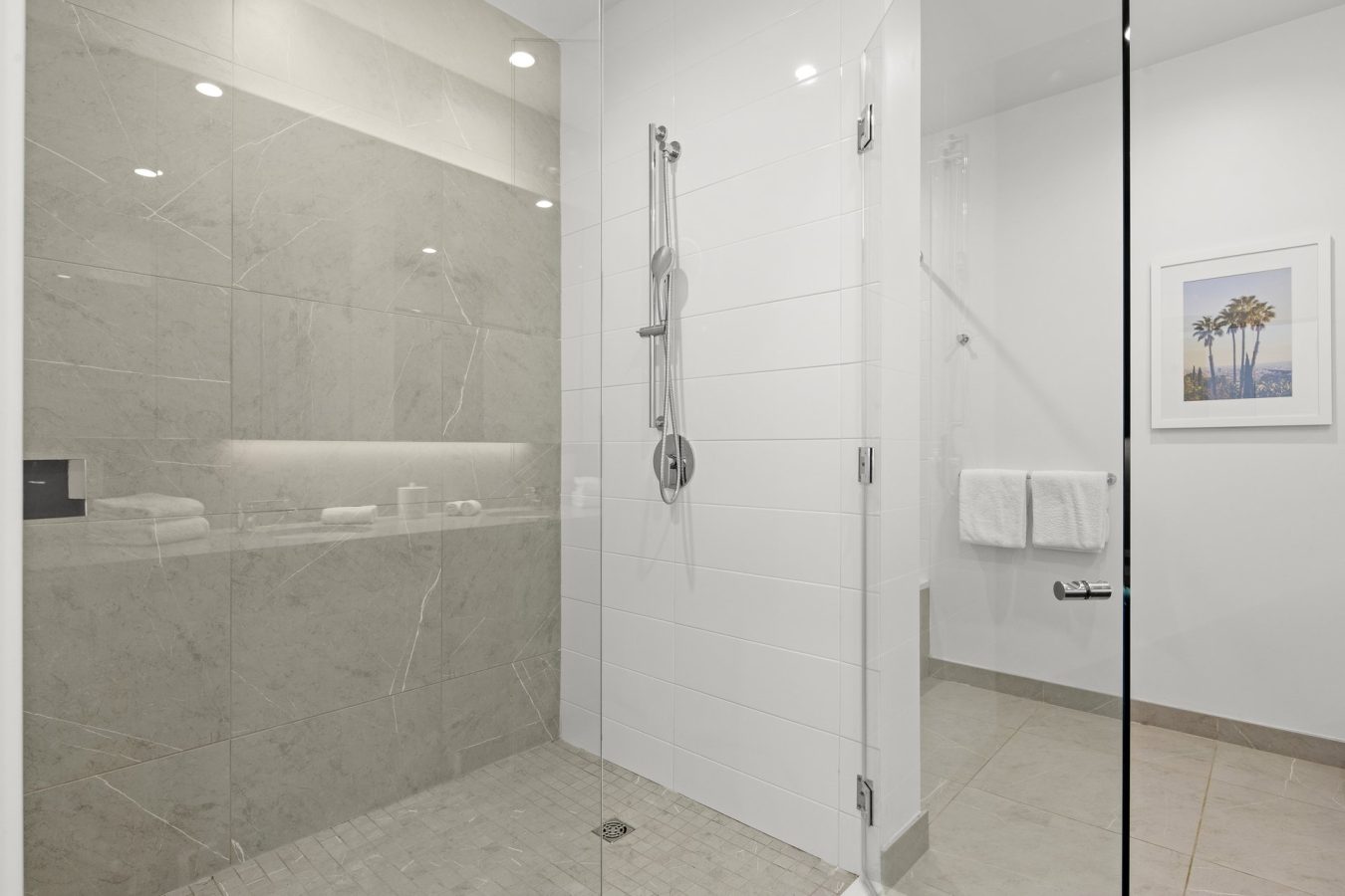 Minimalist bathroom with a glass-enclosed shower featuring beige and white tiles, a chrome showerhead, folded white towels, and a framed picture of palm trees on the wall.