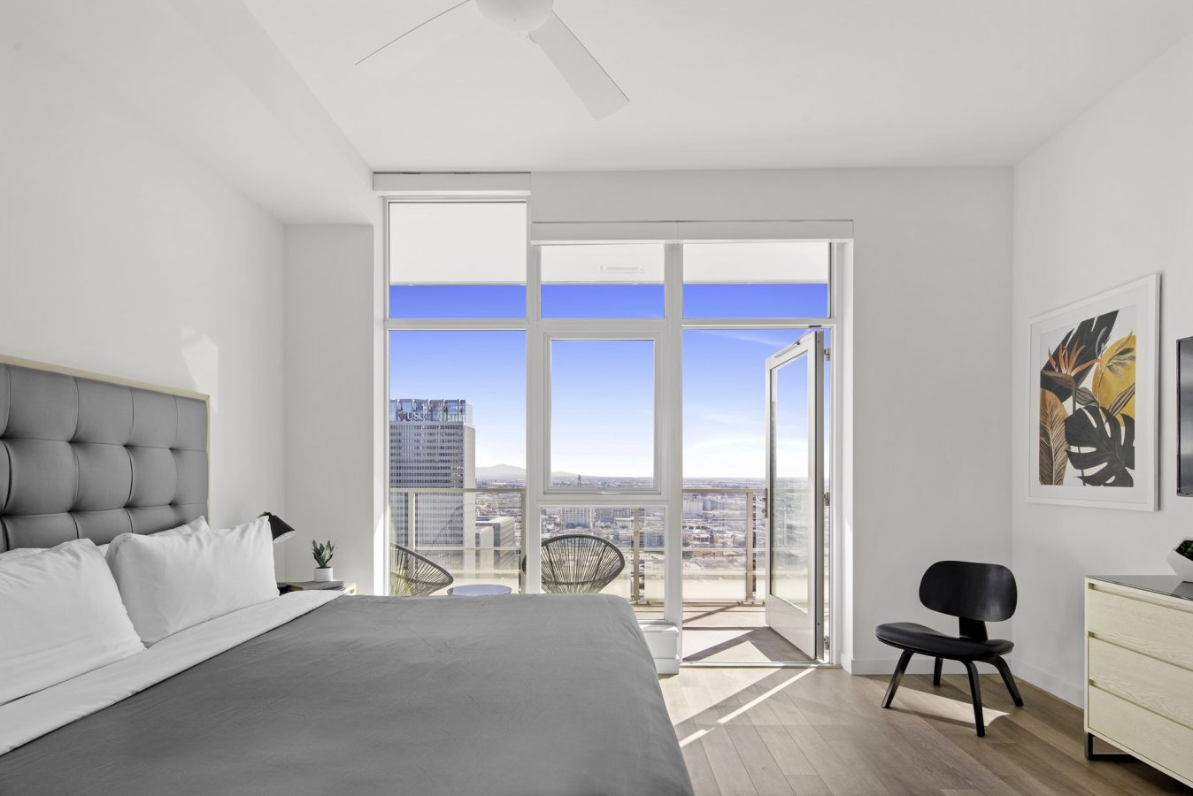 A bright, modern bedroom with a large bed, floor-to-ceiling windows, a balcony with two chairs, a black chair, and minimalist decor, overlooking a cityscape and mountains in the distance.