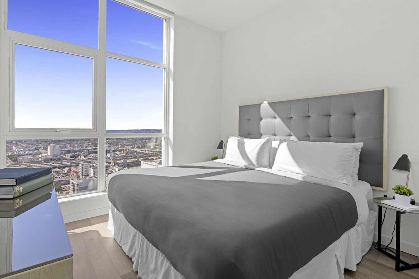 A modern bedroom with a large window offering a city view, a neatly made bed with gray bedding and a padded headboard, bedside lamps, and a desk with books and a small plant.