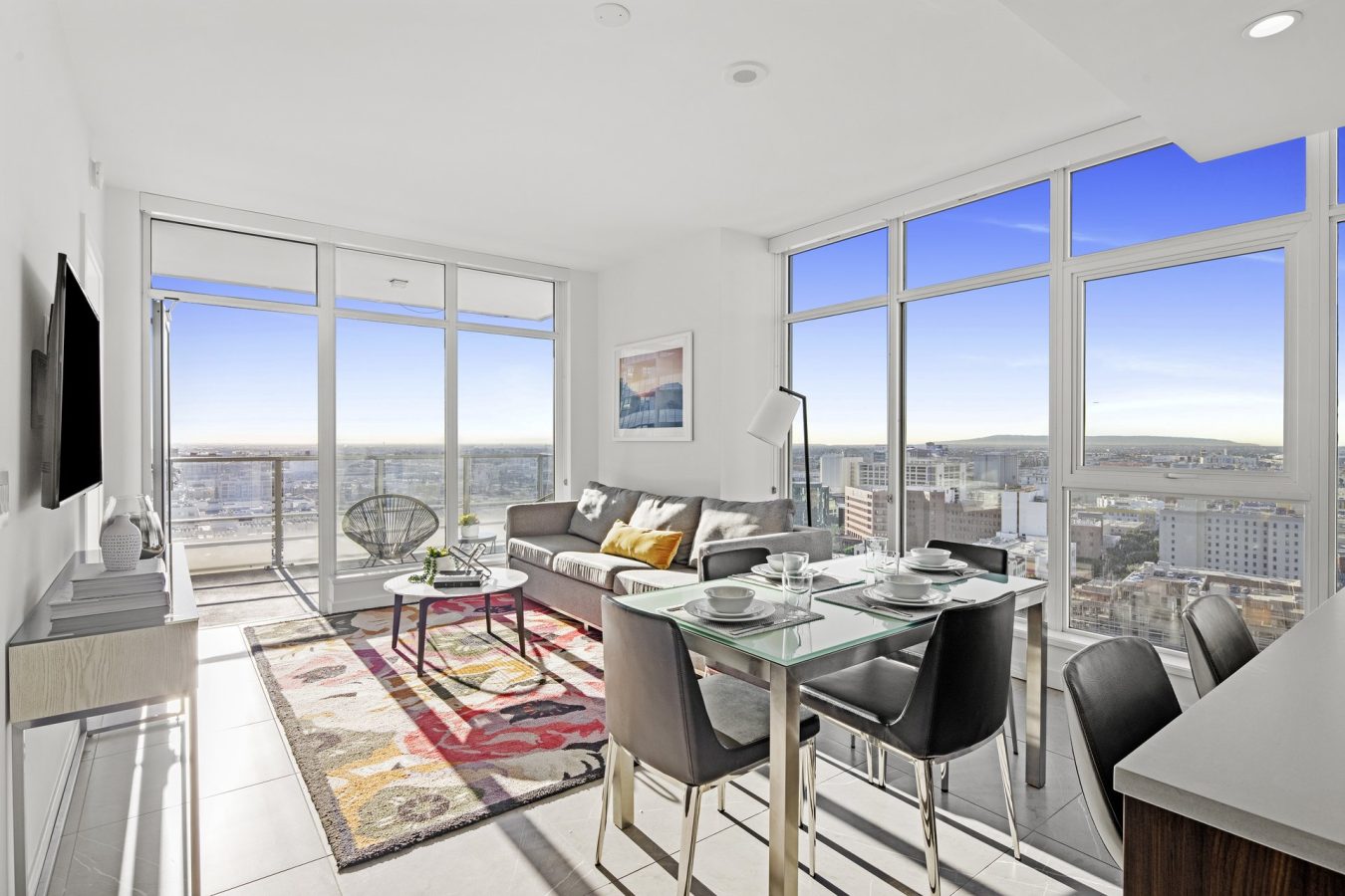Modern, bright apartment living room with large windows, city and ocean views, a gray sofa, colorful rug, glass dining table set for four, and balcony access, decorated in a minimalist style.