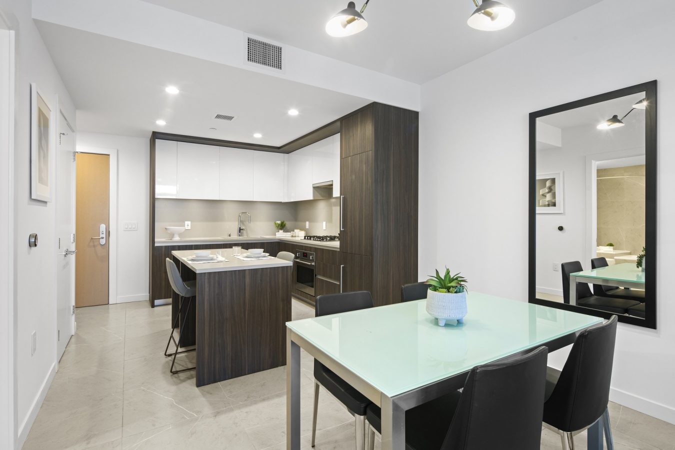 Modern kitchen and dining area with white walls, dark wood cabinets, a small island with bar stools, a glass dining table with four black chairs, and a large wall mirror. A potted plant sits on the table.