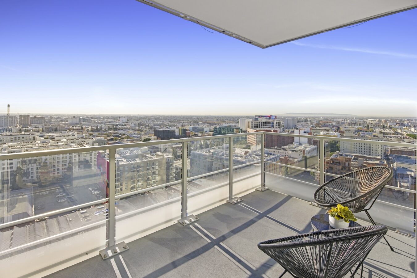A modern balcony with glass railings features two black chairs and a small table with yellow flowers, overlooking a cityscape under a clear blue sky.