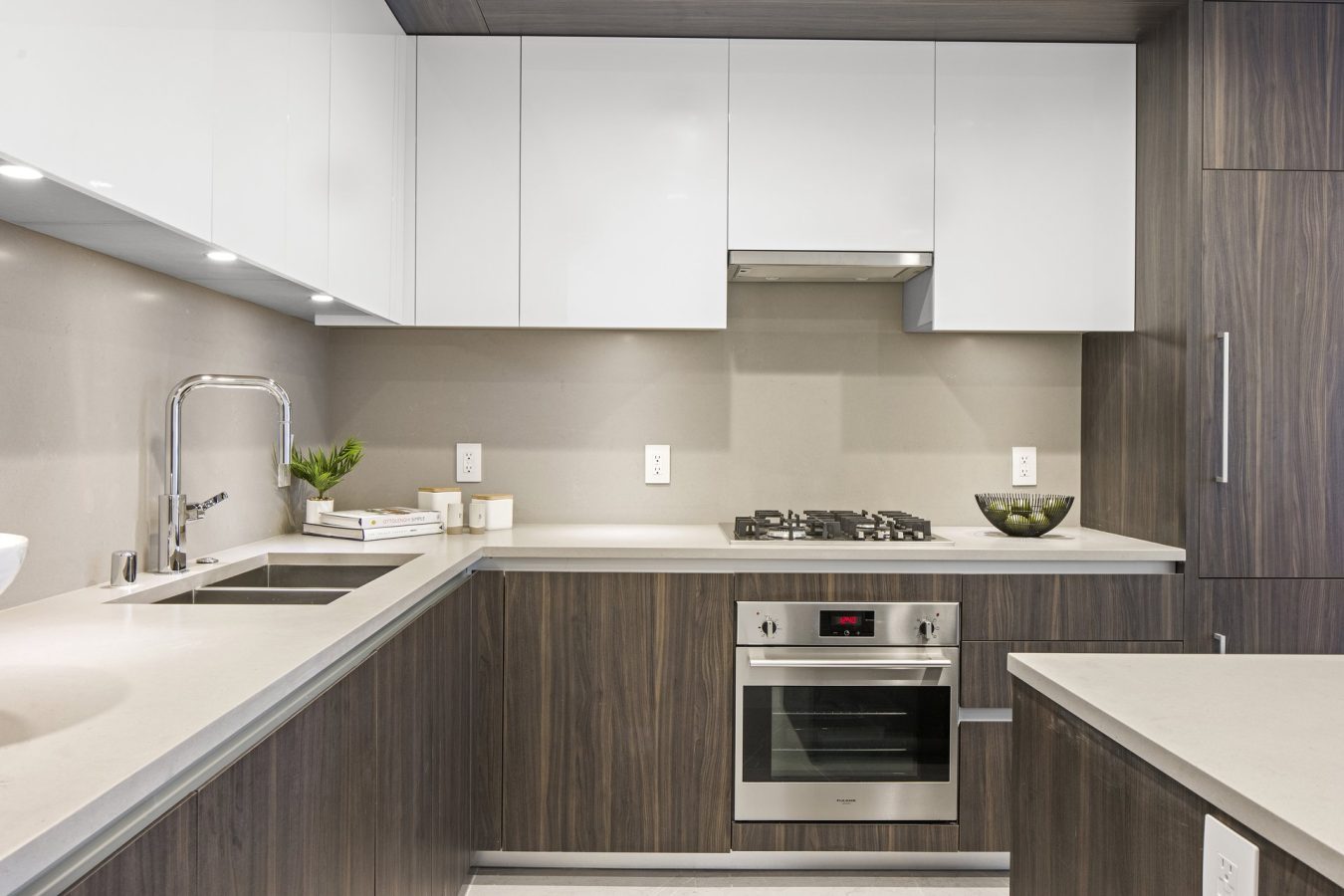 Modern kitchen with brown wood and white cabinets, stainless steel oven, gas stovetop, sink, potted plant, stacked books, and a bowl of green apples on light-colored countertops.