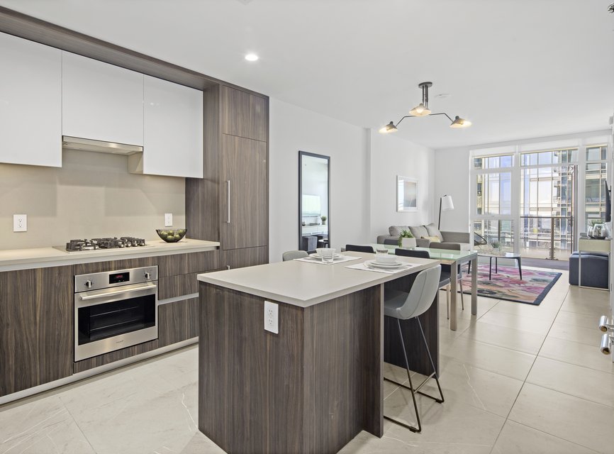 Modern apartment kitchen with wood cabinetry, stainless steel appliances, and a center island with two stools. The open living area includes a sofa, dining table, large window, and contemporary lighting.