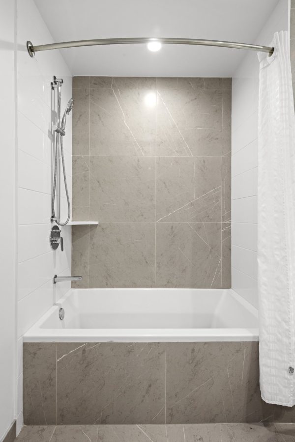 Modern bathroom with a white bathtub, beige marble-style tile walls, a mounted showerhead, a small wall shelf, and a curved shower rod with a white shower curtain pulled to one side.