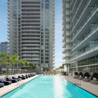 A long outdoor swimming pool is surrounded by lounge chairs and modern high-rise apartment buildings, with palm trees lining the poolside under a clear blue sky.