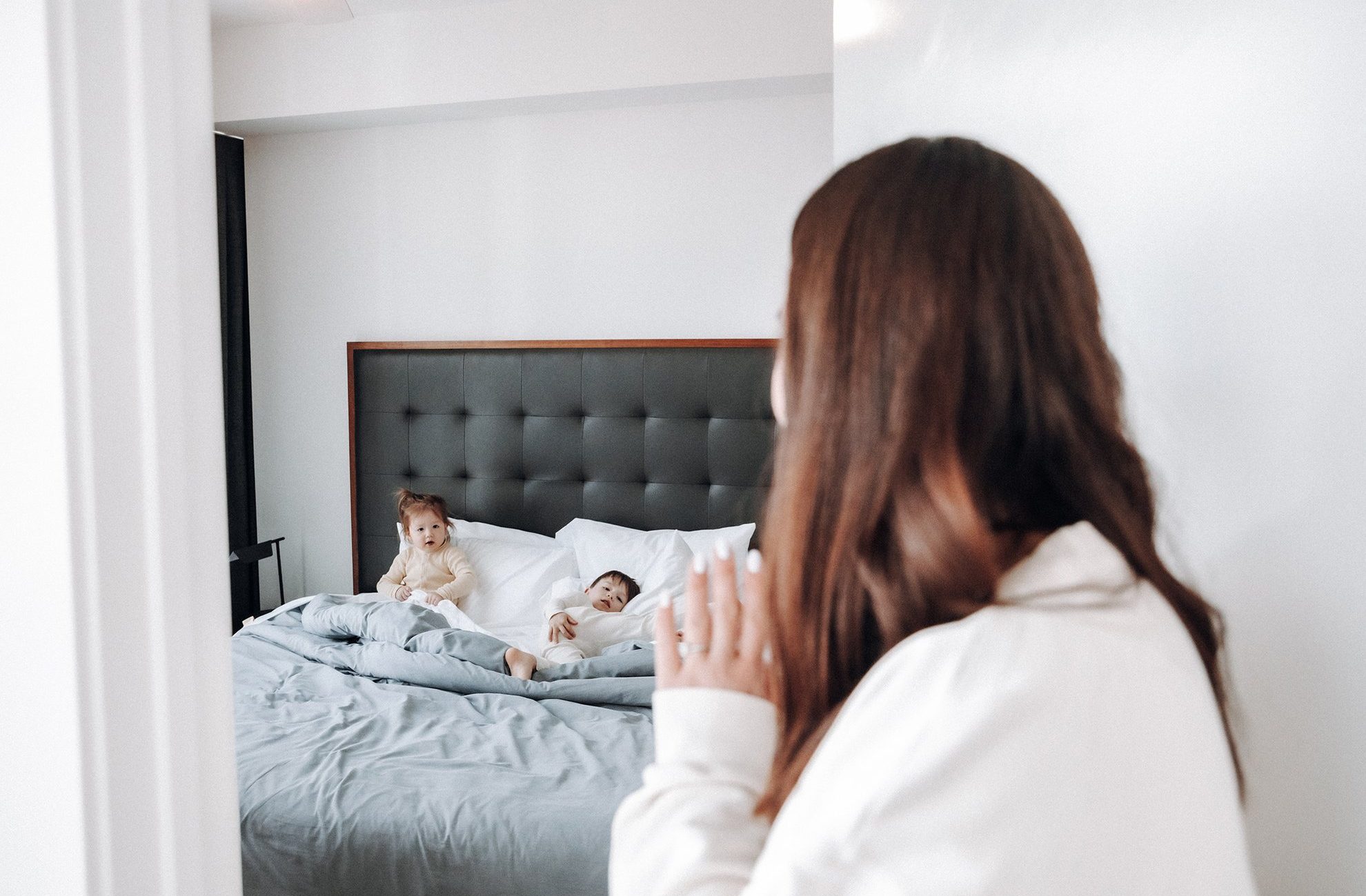 A woman with long brown hair stands in a doorway, facing two young children sitting on a bed with grey bedding in a bright, modern bedroom. The children are looking toward the woman.