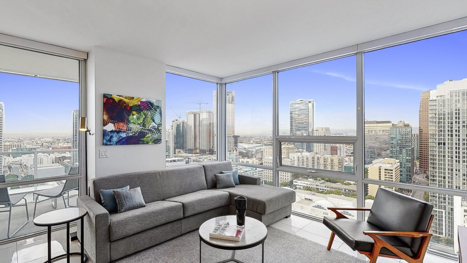 Modern living room with large windows offering a panoramic city skyline view, featuring a gray sofa, black armchair, abstract wall art, and a round coffee table with magazines and a cup.