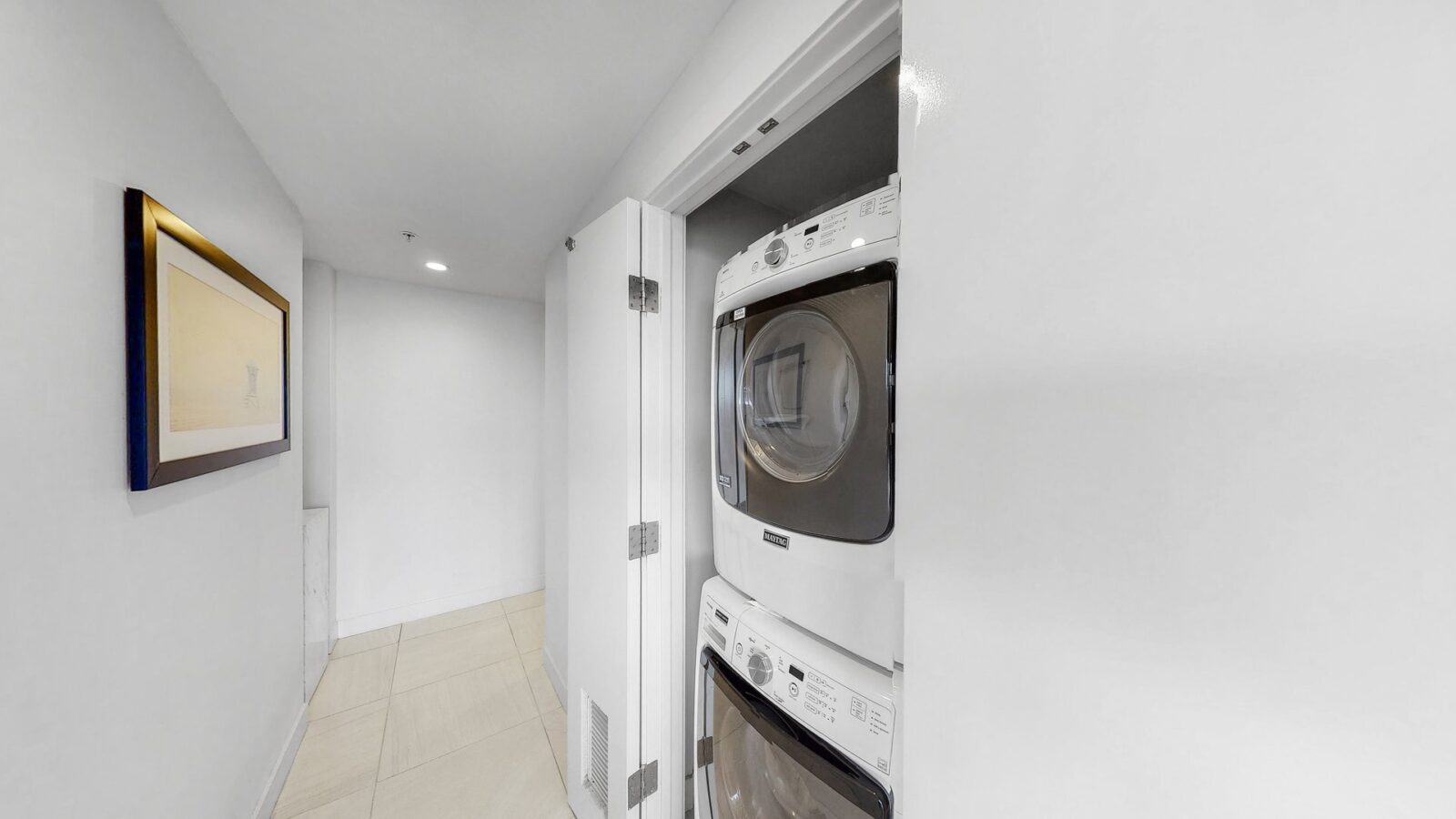 A bright, modern laundry area with stacked front-loading washer and dryer behind a partially open white door, next to a white hallway with a framed picture on the wall.