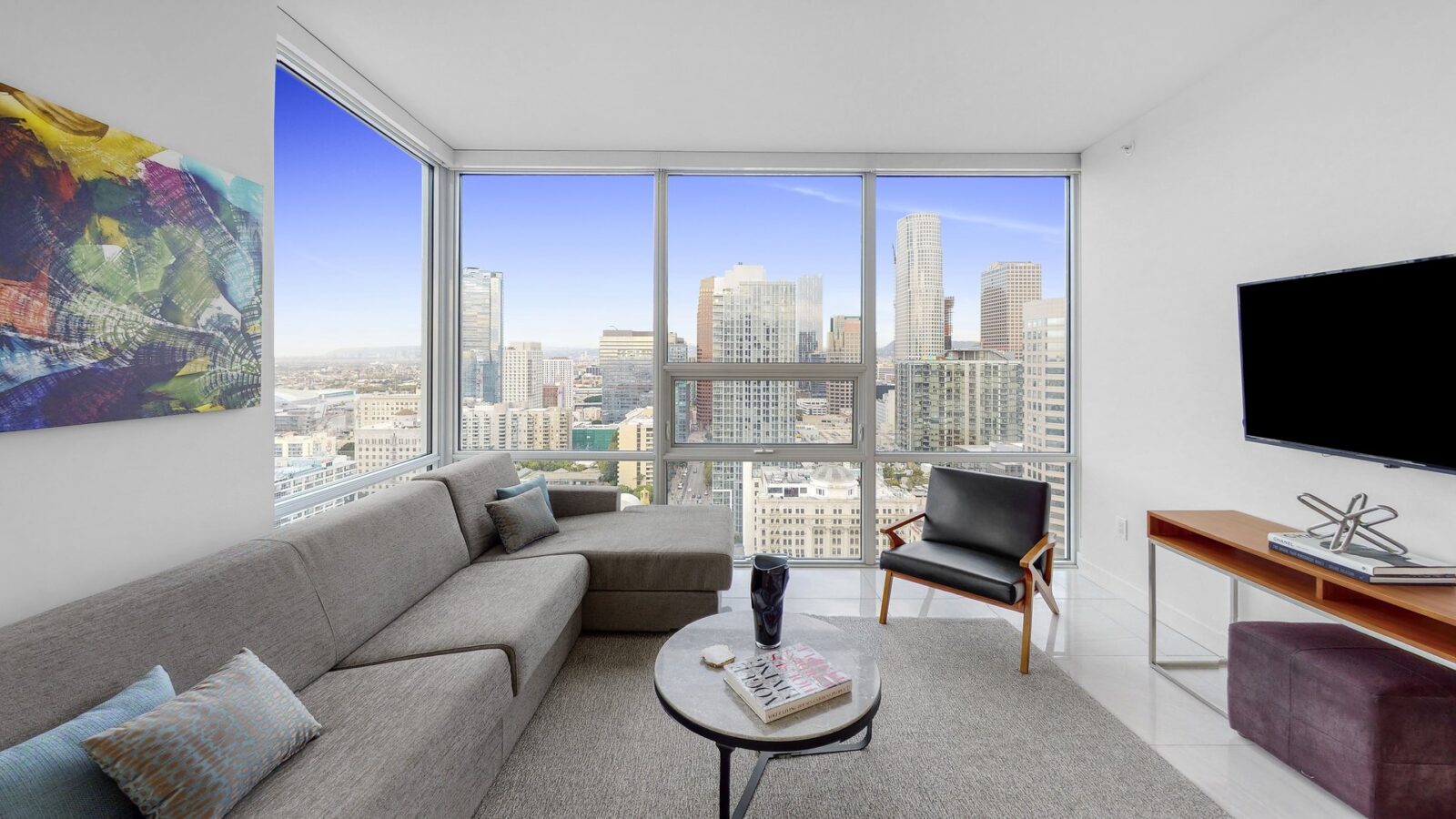 Modern living room with a gray sectional sofa, armchair, and coffee table, featuring floor-to-ceiling windows with a view of a city skyline. Abstract art and a wall-mounted TV decorate the white walls.