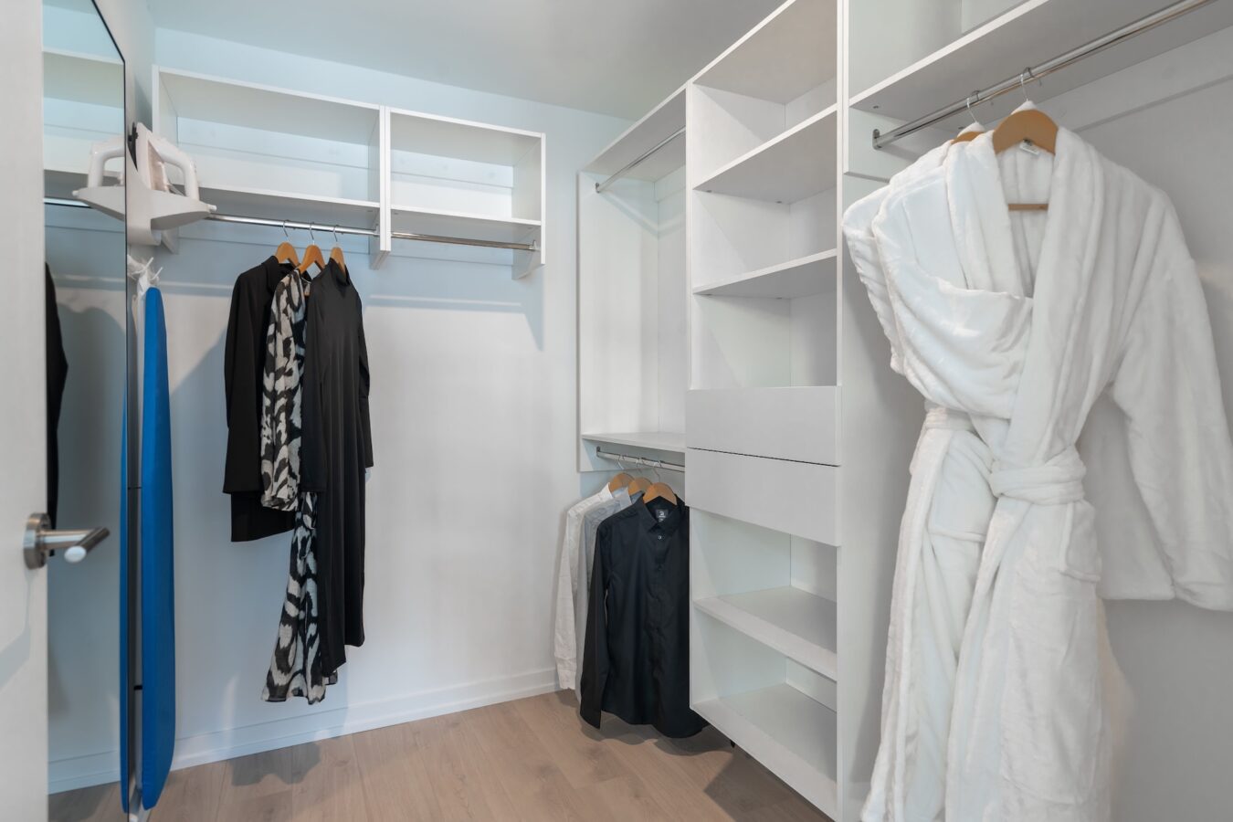 A modern walk-in closet with white shelves and rods, a hanging white robe, several black and white clothes on wooden hangers, a blue ironing board, and light wood flooring.