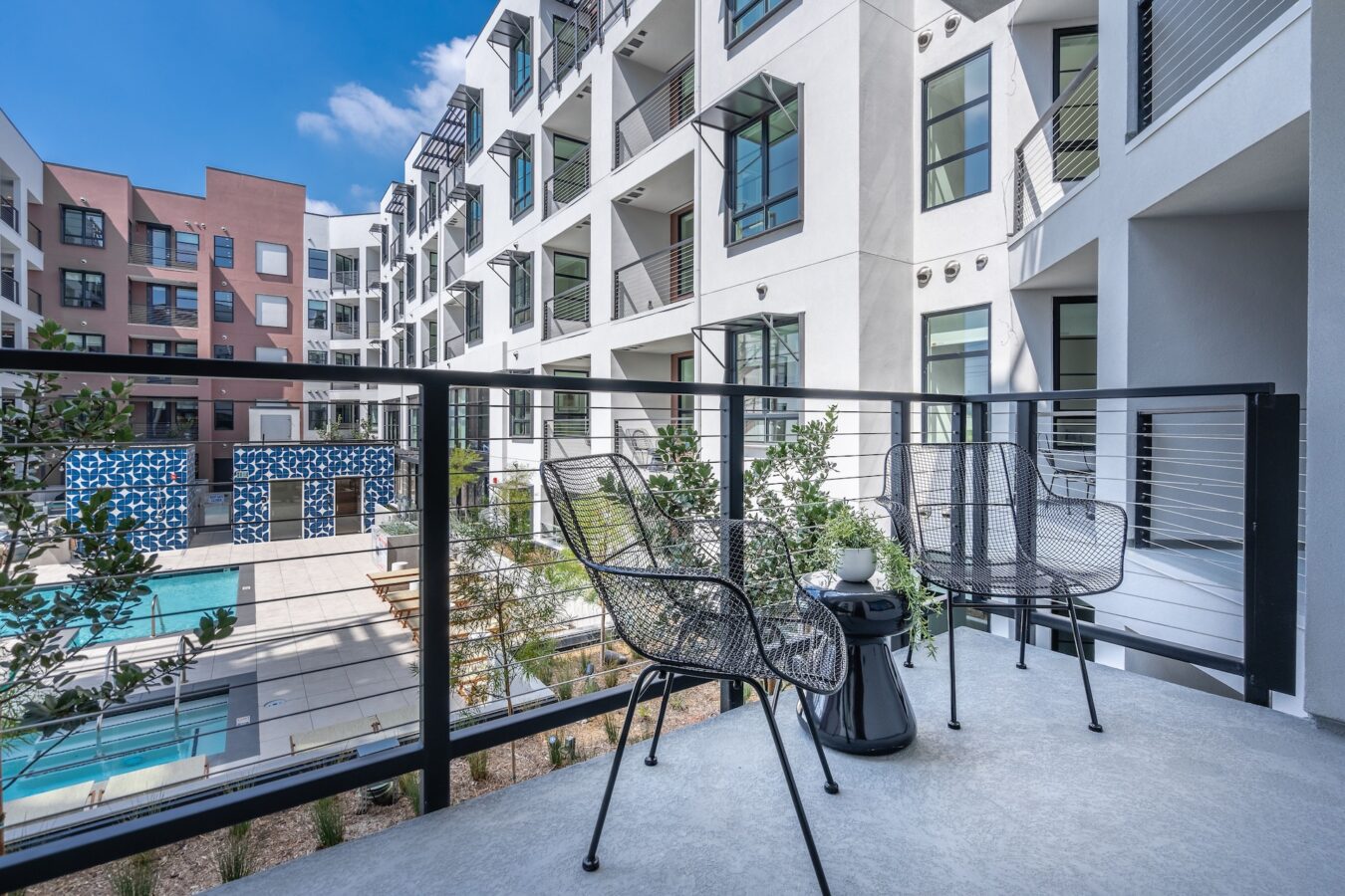 A modern apartment balcony with two black mesh chairs and a small table overlooks a courtyard with a swimming pool, landscaping, and neighboring apartment buildings under a clear blue sky.