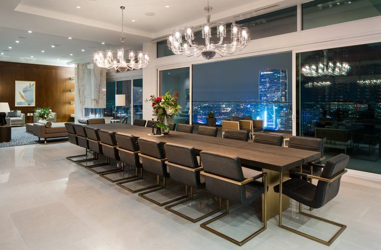 Modern dining room with a long dark wood table, black chairs, two large chandeliers, floor-to-ceiling windows, city skyline view at night, and a stylish living area in the background.