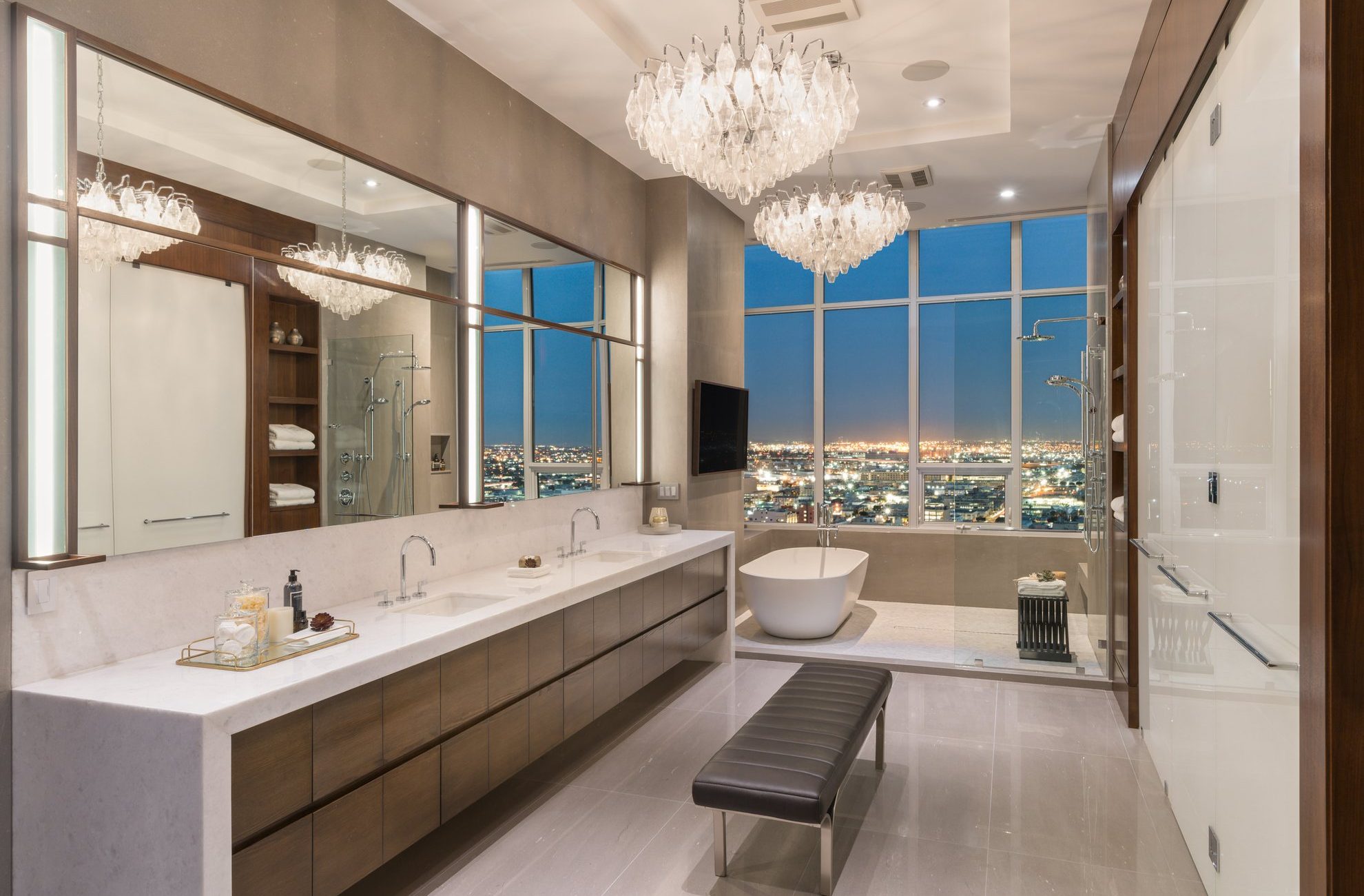 Modern luxury bathroom with double sinks, a large mirror, and glass shower. A freestanding tub sits by floor-to-ceiling windows overlooking a city at night. Elegant chandeliers hang from the ceiling, and a bench is in the center.