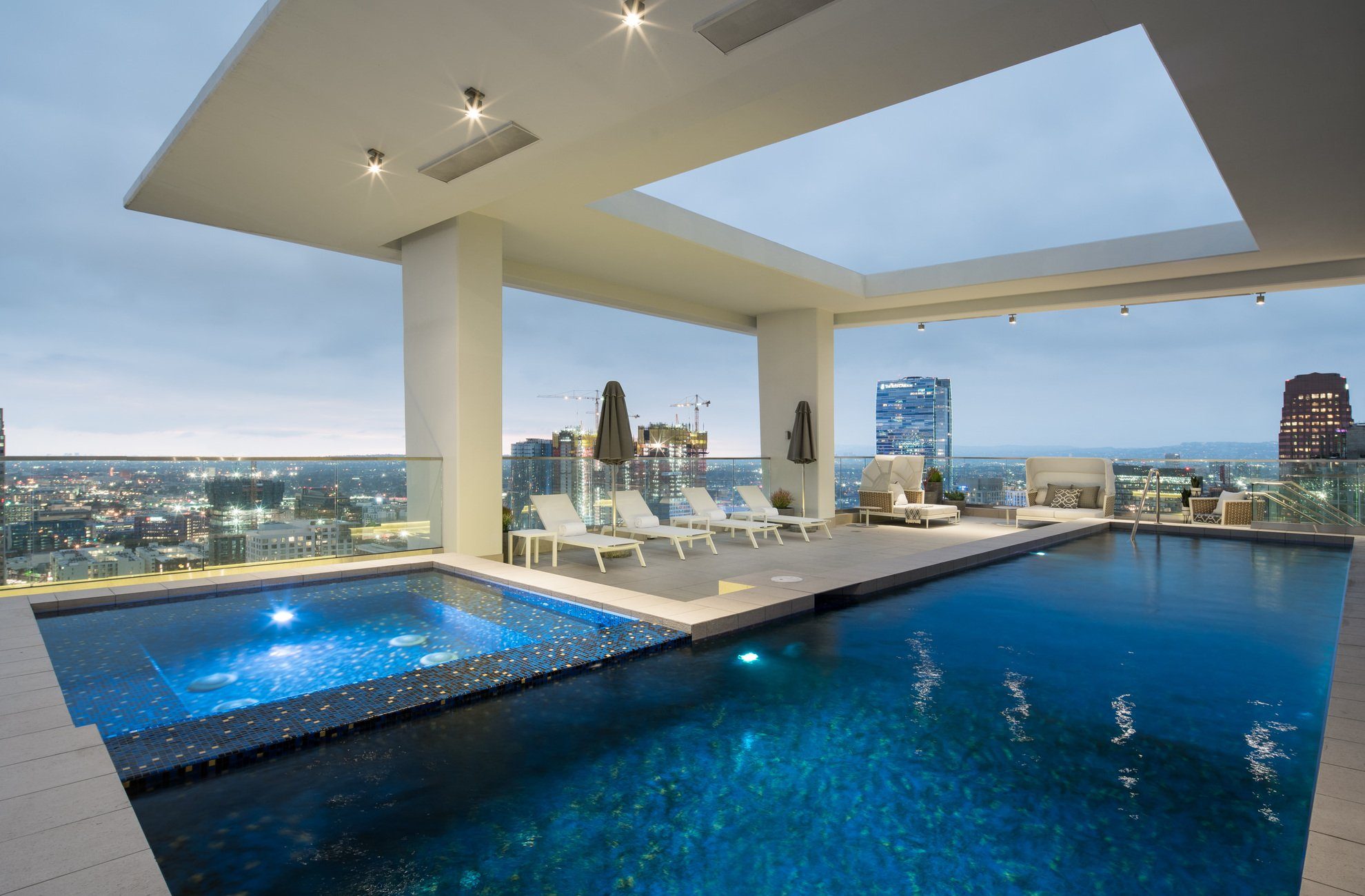 A modern rooftop pool with lounge chairs, umbrellas, and city skyscrapers visible in the background at dusk. The pool area is covered by a large open ceiling structure with built-in lights.