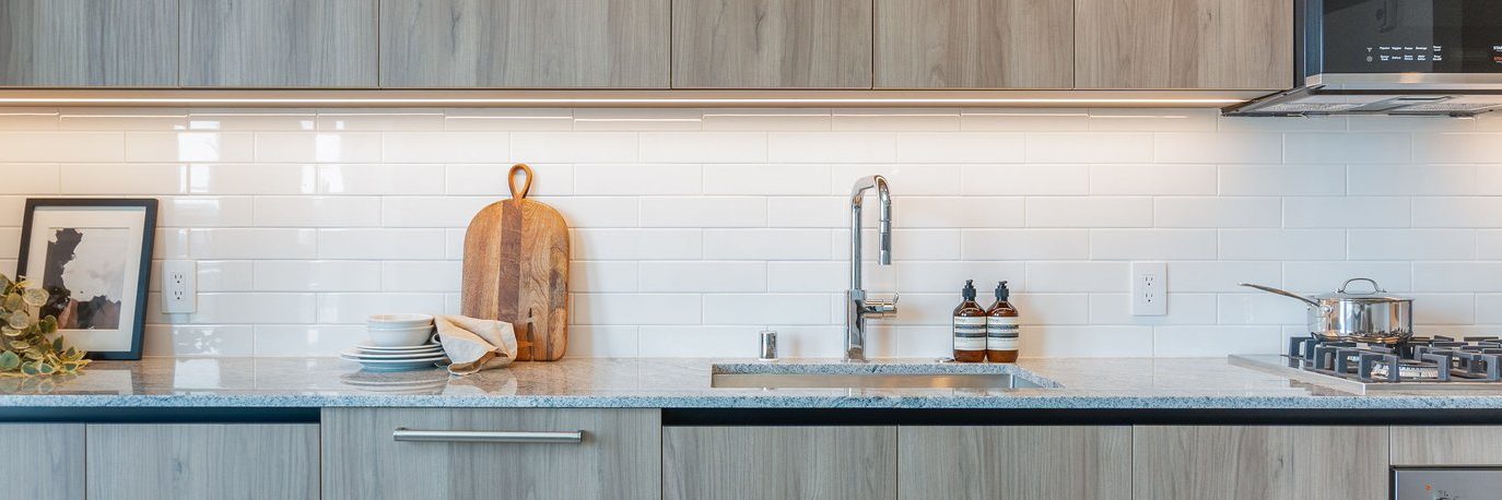 Modern kitchen with light wood cabinets, gray countertop, white subway tile backsplash, a sink with a tall faucet, a cutting board, stacked dishes, soap bottles, and a stove with a pot.