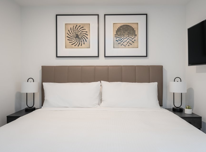 A neatly made bed with a brown tufted headboard, white bedding, and two white pillows. Two modern lamps sit on black nightstands, and above the bed hang two framed abstract art pieces. A TV is mounted on the right wall.