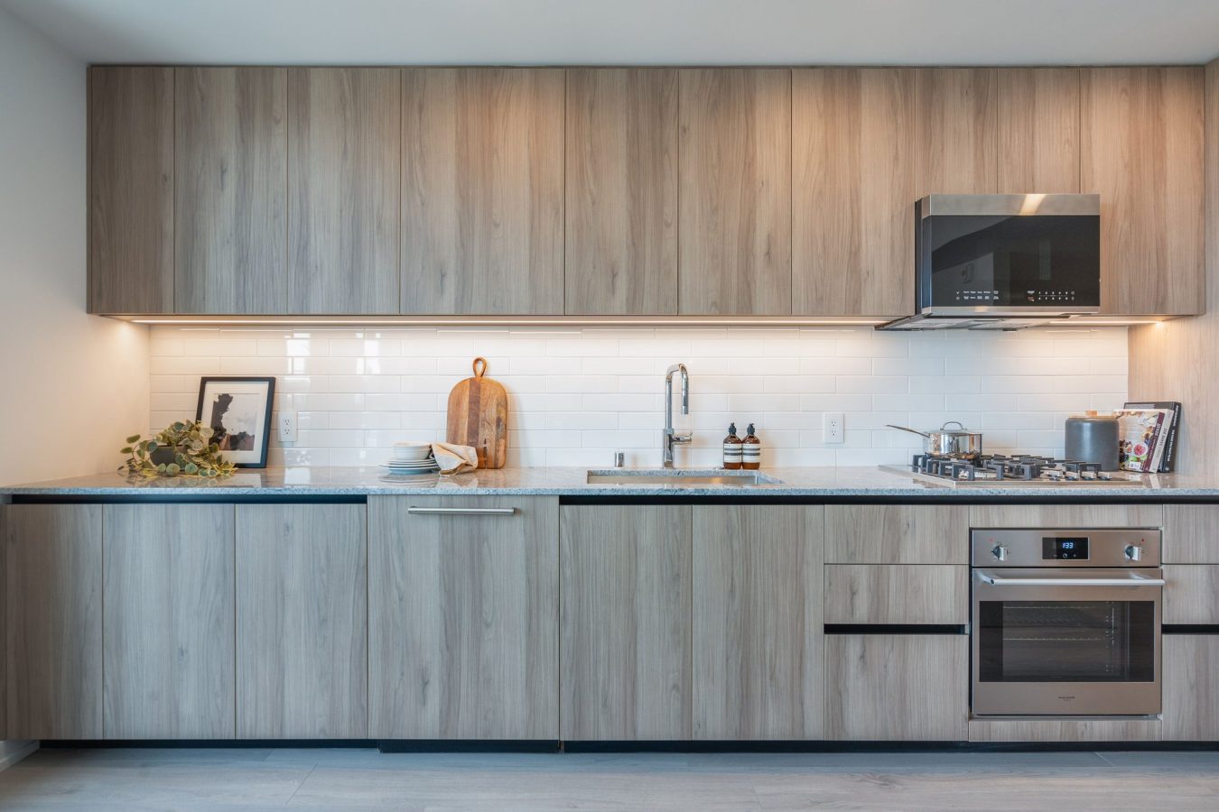 Modern kitchen with light wood cabinets, white subway tile backsplash, built-in oven, gas stove, and stainless steel range hood. Countertop has a cutting board, framed photo, dishes, and small decor items.