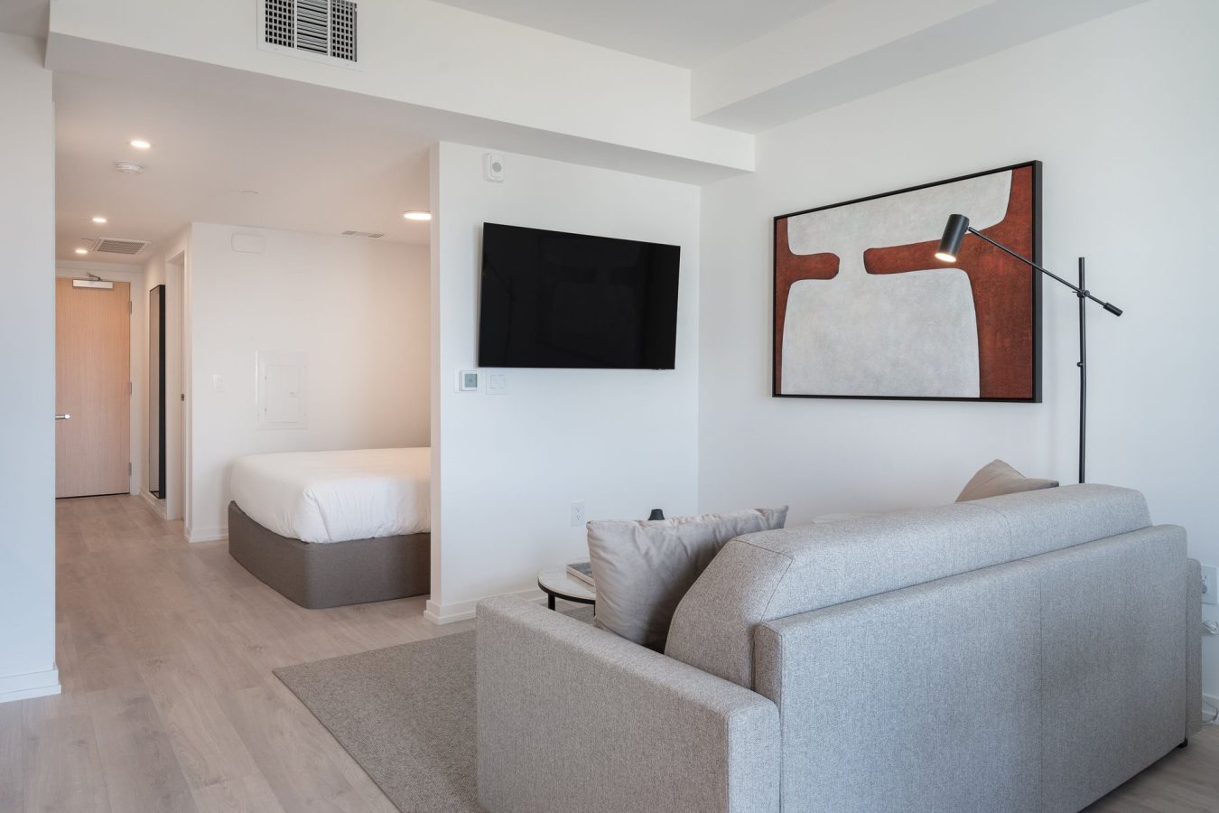A modern, minimalistic studio apartment with a gray sofa, wall-mounted TV, abstract artwork, floor lamp, and a visible bed area through an open doorway. Light wood floors and white walls give the space an airy feel.