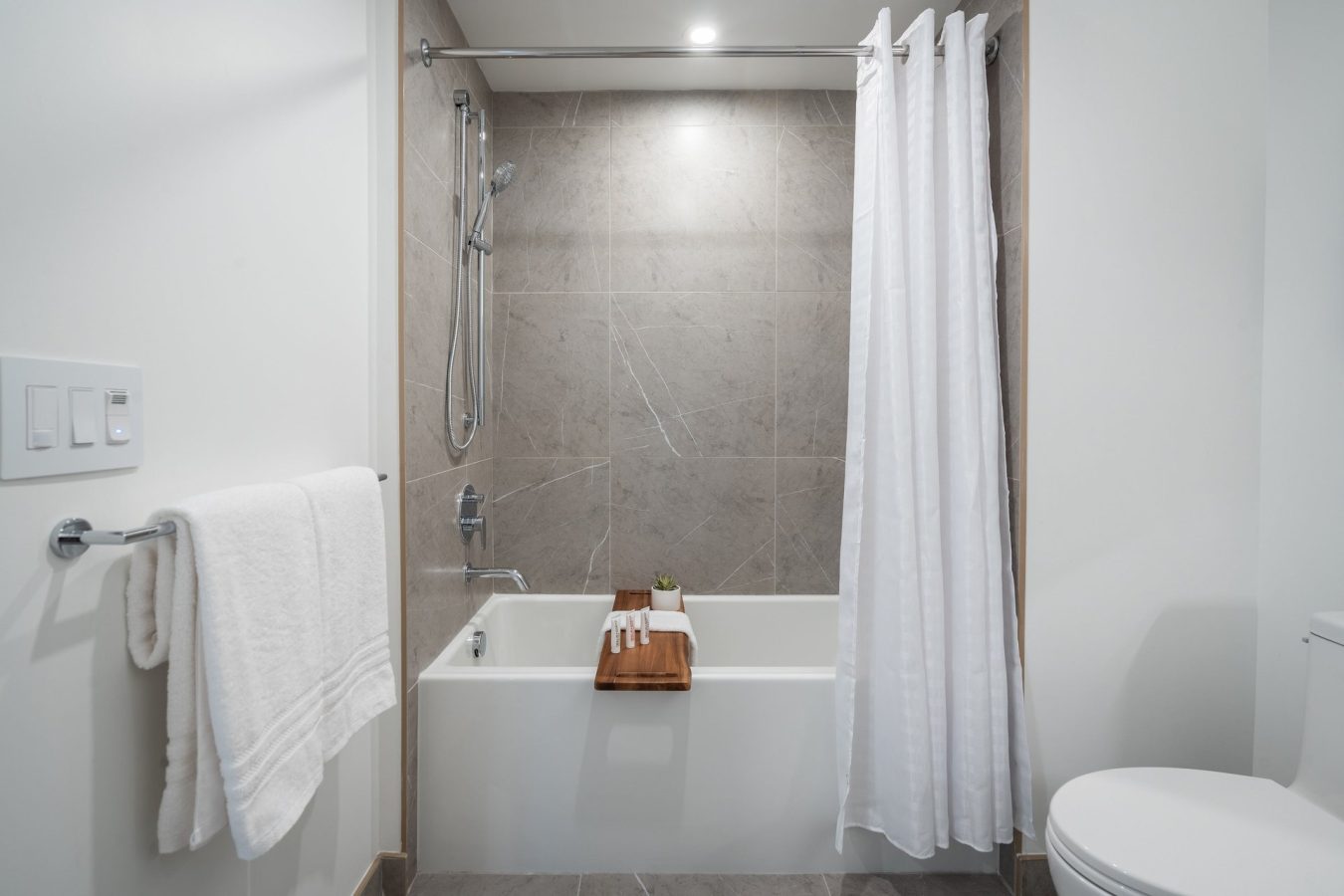 Modern bathroom with a white bathtub, gray tiled walls, a showerhead, and a white shower curtain. A wooden tray with towels and a small plant rests on the tub. White towels hang on a bar nearby.
