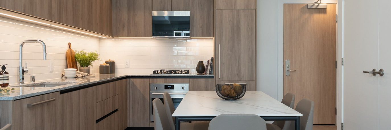 Modern kitchen with wood cabinets, built-in appliances, and marble dining table with four chairs. White tile backsplash, under-cabinet lighting, and a bowl of fruit on the table. Door leading to another room is visible.