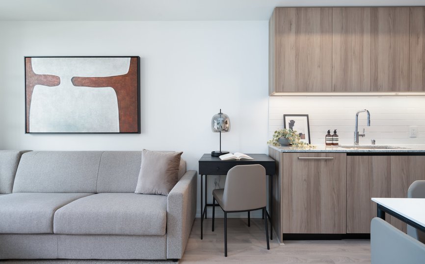 A modern living space with a light gray sofa, abstract wall art, a small black desk and beige chair, and a kitchenette with wood cabinets and a white countertop. The space is bright and minimalistic.