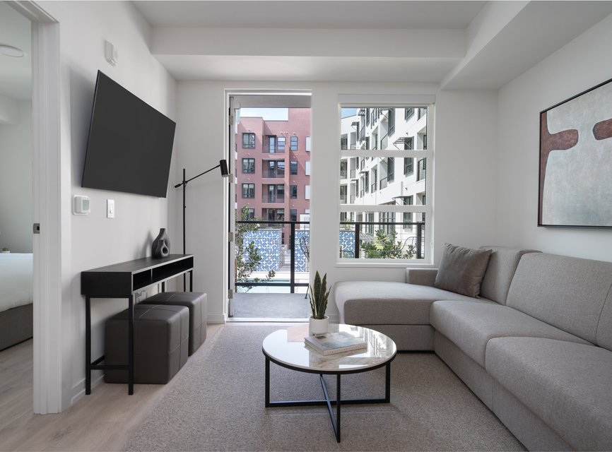 Modern living room with a gray sectional sofa, round coffee table, wall-mounted TV, floor lamp, and large windows leading to a balcony overlooking neighboring apartment buildings. Neutral decor and contemporary art on the wall.