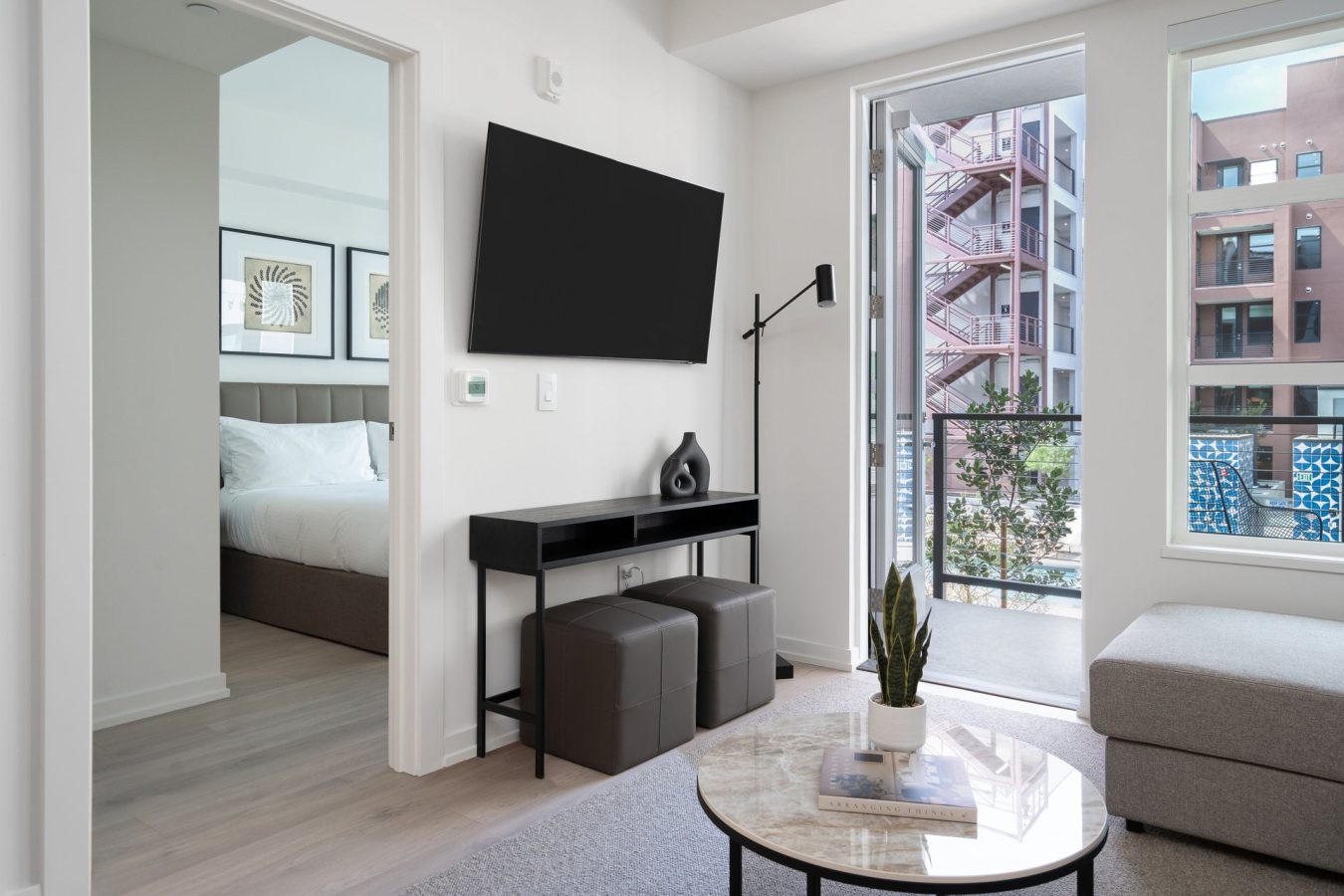 Modern apartment living room with a wall-mounted TV, console table, two ottomans, and a round coffee table with a plant; bedroom and balcony with city view are visible in the background.