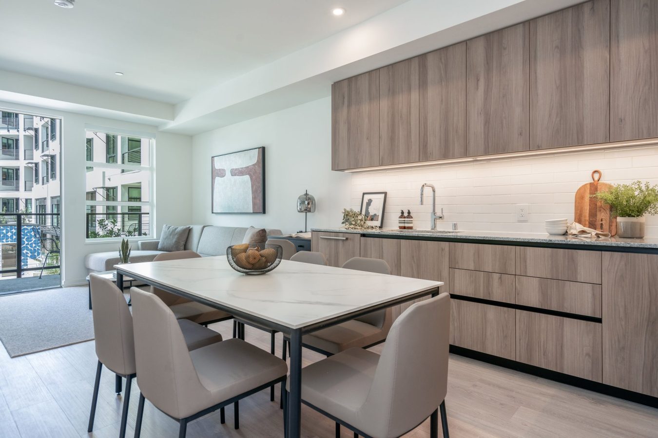 Modern apartment kitchen and dining area with light wood cabinets, marble-top dining table, beige chairs, a cozy sofa, large windows, and minimalist decor, creating a bright and inviting open living space.