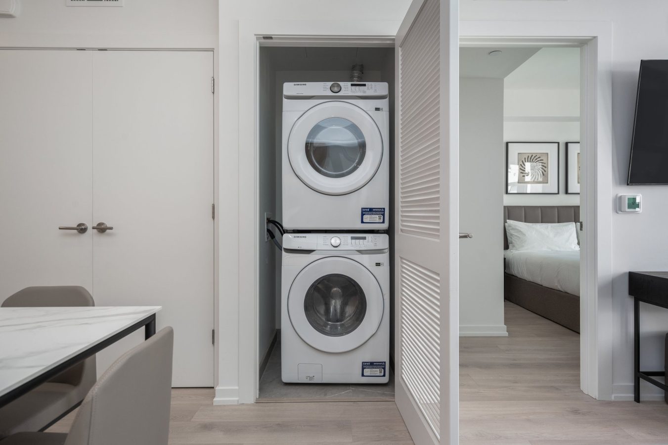 A stacked washer and dryer are neatly placed in a small laundry closet with a louvered door, between a dining area and a bedroom in a modern, light-colored apartment.