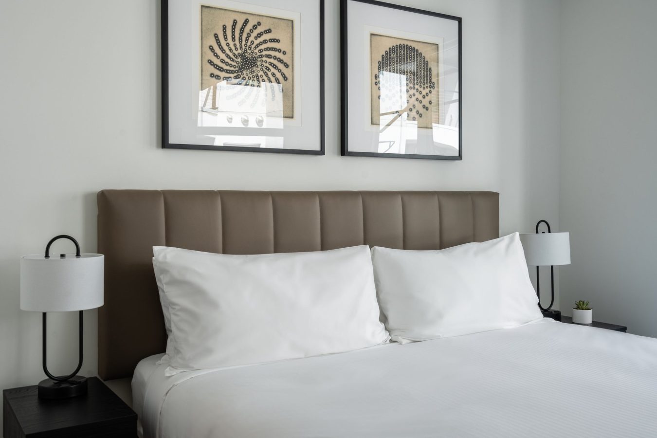 A neatly made bed with white linens and a brown padded headboard, flanked by two black nightstands with modern white lamps. Two framed abstract artworks hang on the light-colored wall above the bed.