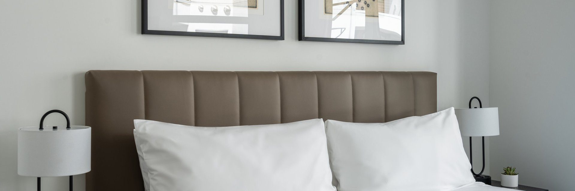 A neatly made bed with white pillows and a brown upholstered headboard, flanked by two modern bedside lamps. Two framed artworks hang on the light-colored wall above the bed.